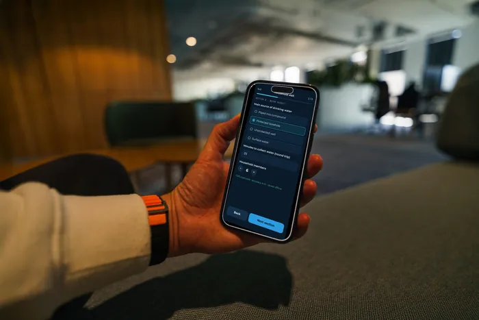 A hand holding a phone showing a household survey form with sample data.